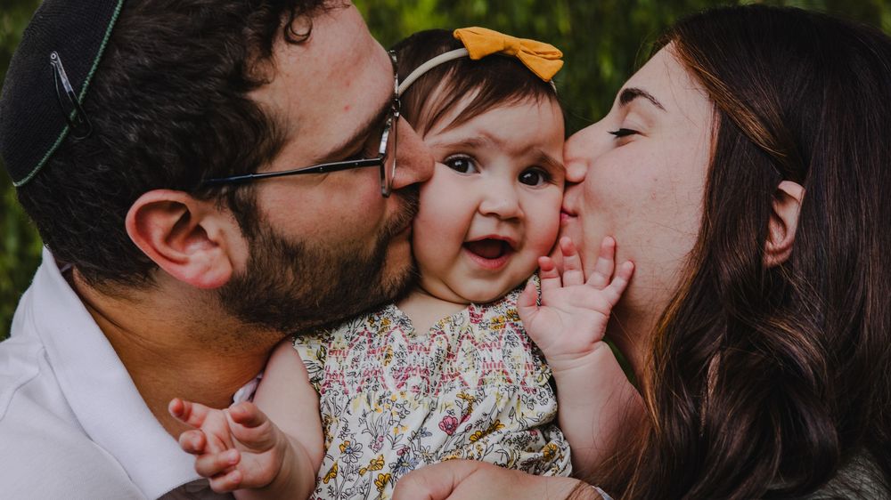 Parents kissing their smiling baby girl wearing a floral dress and yellow bow headband outdoors.