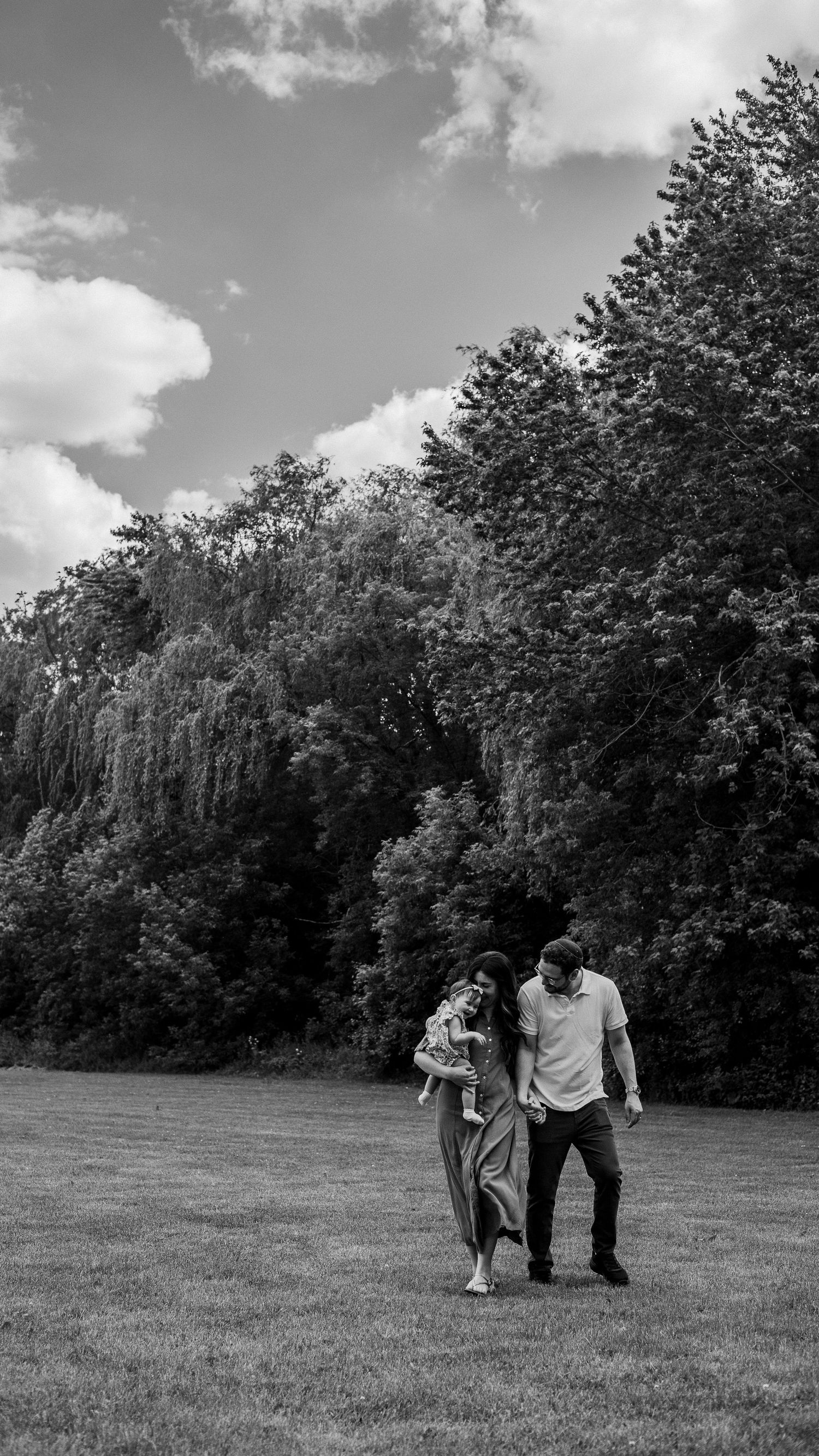 Black and white photo of a couple walking together on a grassy field surrounded by lush trees under a cloudy sky.