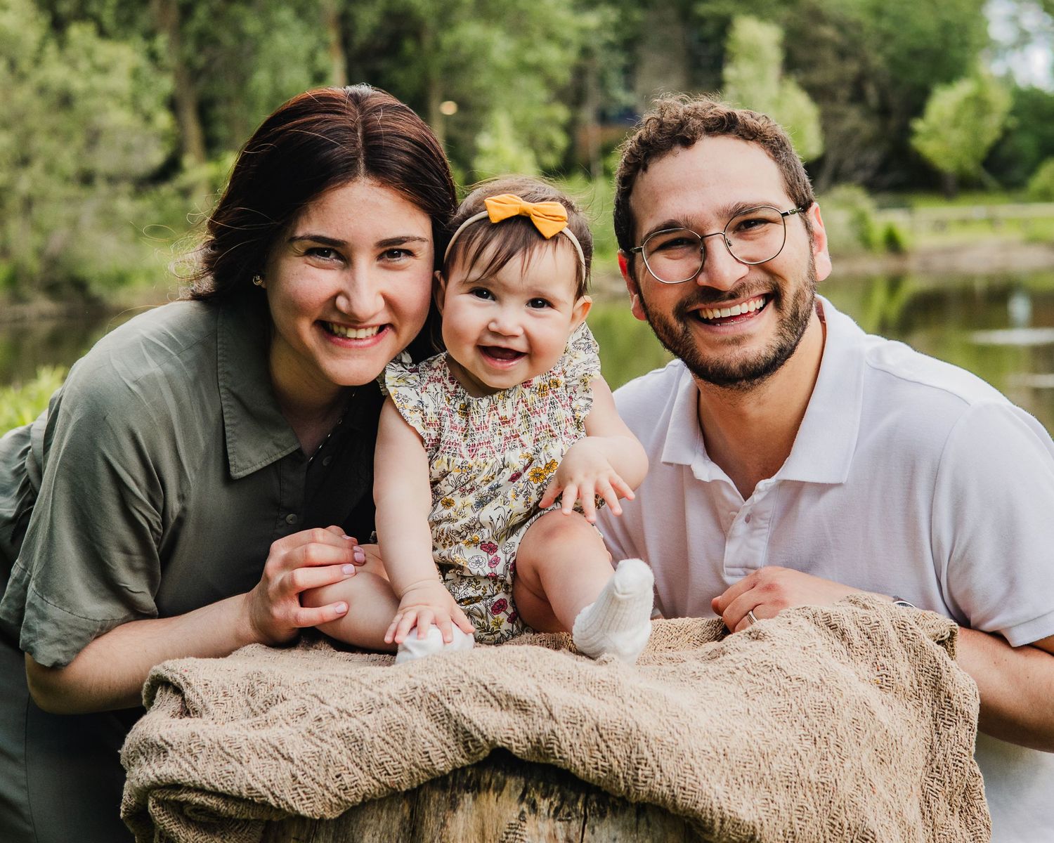 Happy young family with smiling parents and adorable baby girl posing outdoors near a pond in a lush green park.