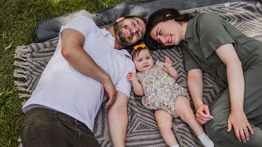Family of three lying on a blanket in the grass, parents smiling beside their baby girl in a floral outfit.