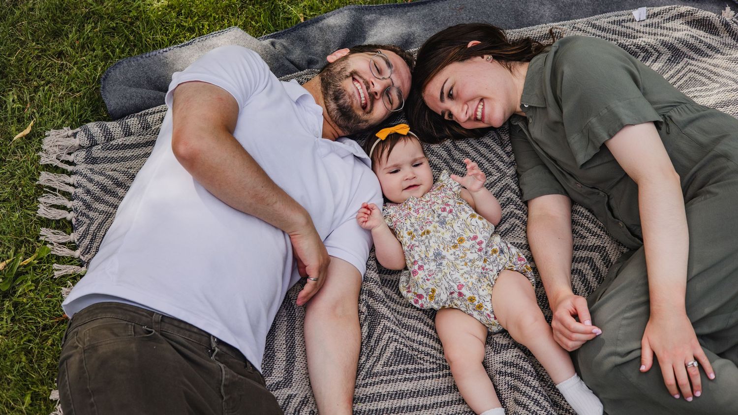 Family of three lying on a blanket in the grass, parents smiling beside their baby girl in a floral outfit.