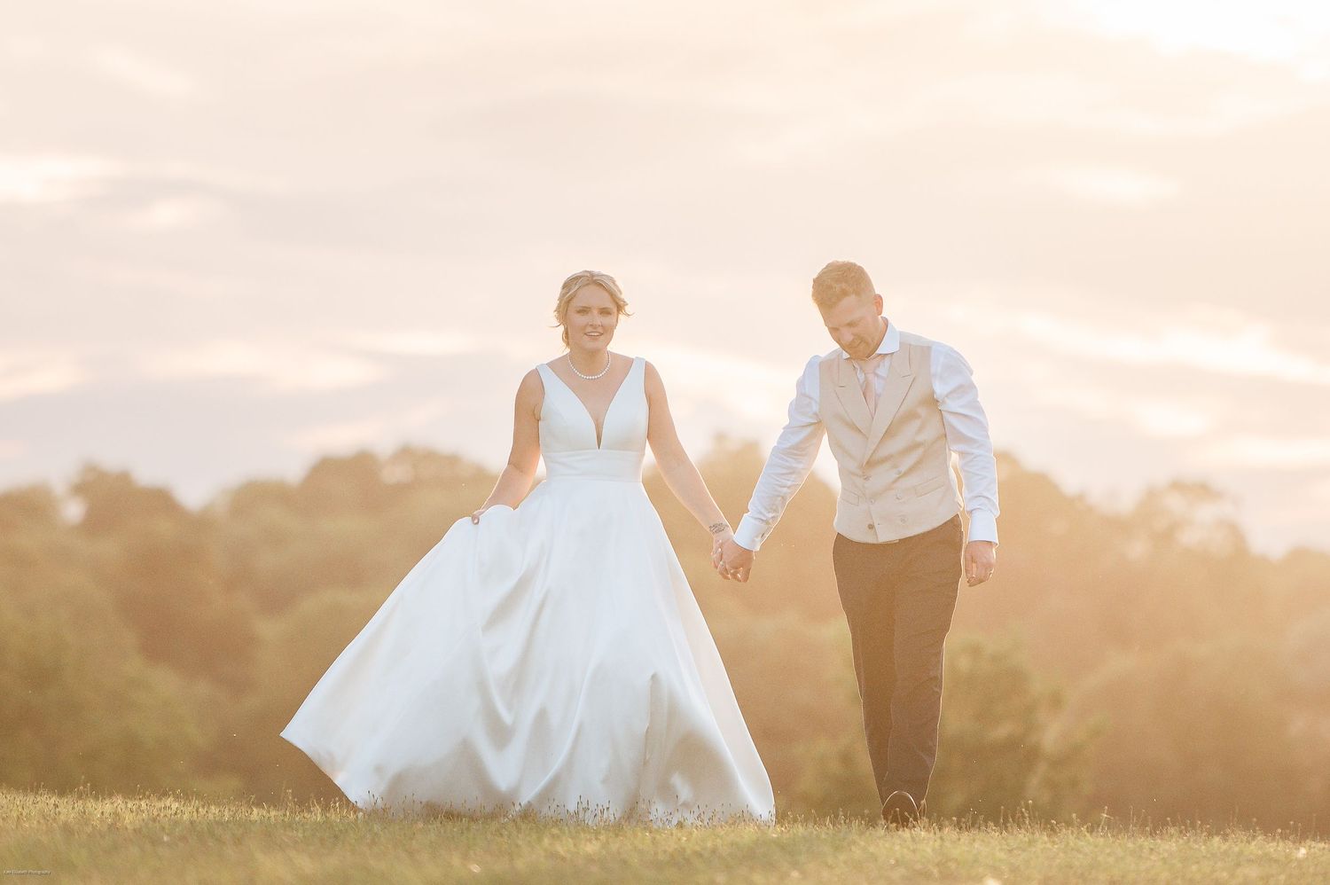 A couple in wedding attire holding hands while walking through a sunlit meadow at golden hour.