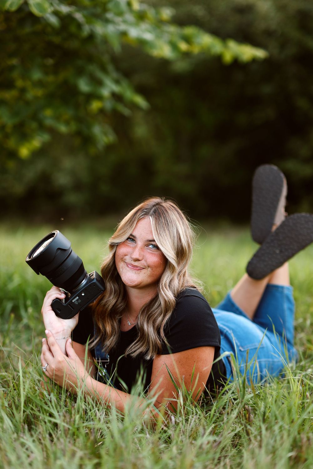 Professional photographer in gray off-shoulder sweater and jeans holds camera while posing against white background.