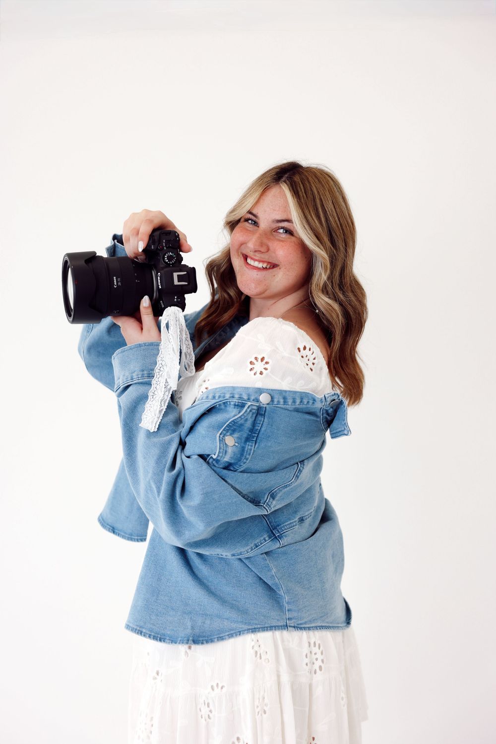 Professional photographer in gray off-shoulder sweater and jeans holds camera while posing against white background.