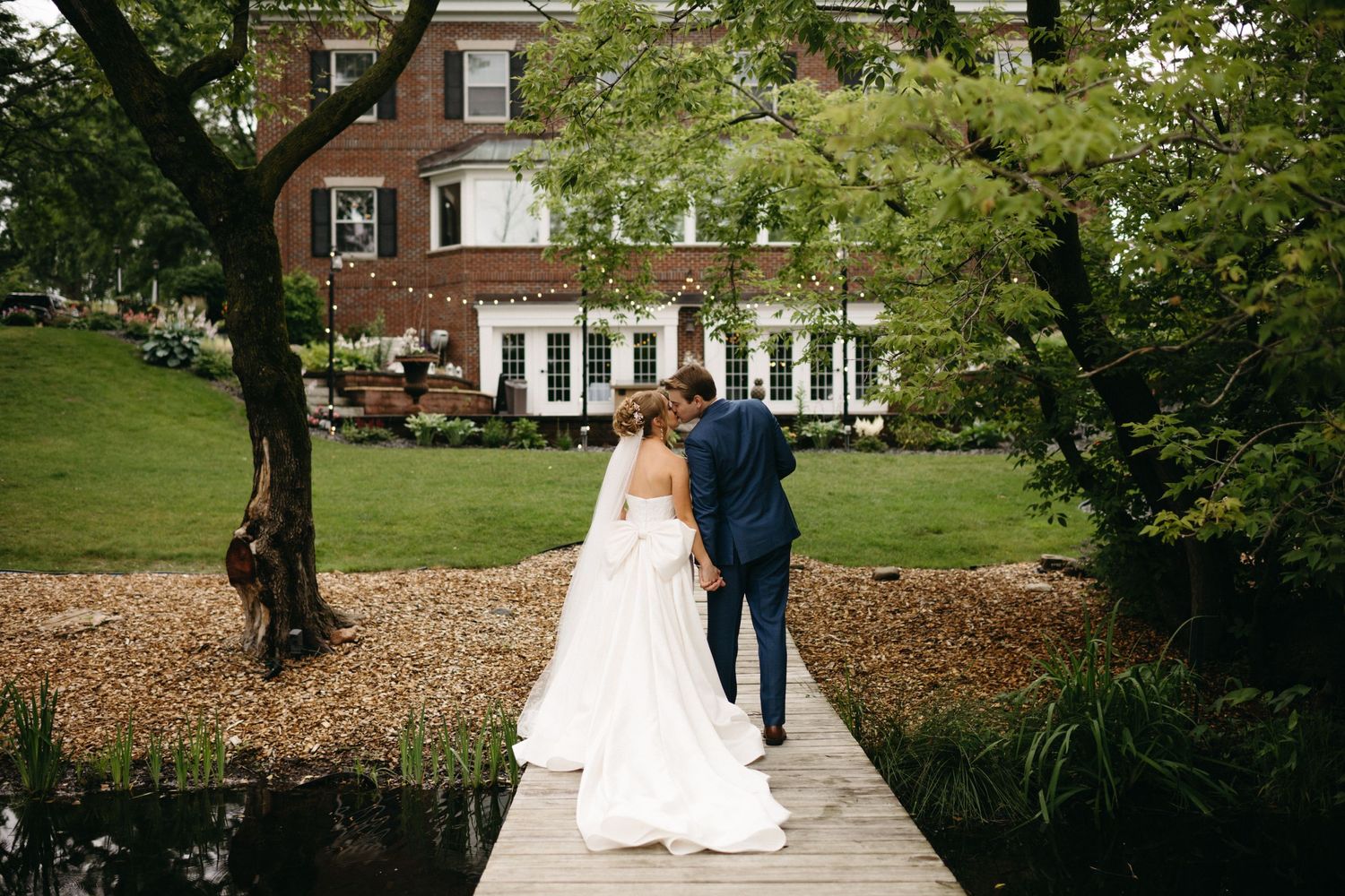 Wedding couple walking along stone path towards brick mansion surrounded by lush gardens.