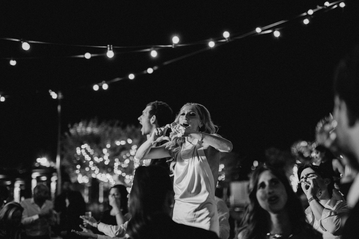 A black and white photo of performers on stage at night beneath glowing string lights during an outdoor concert.