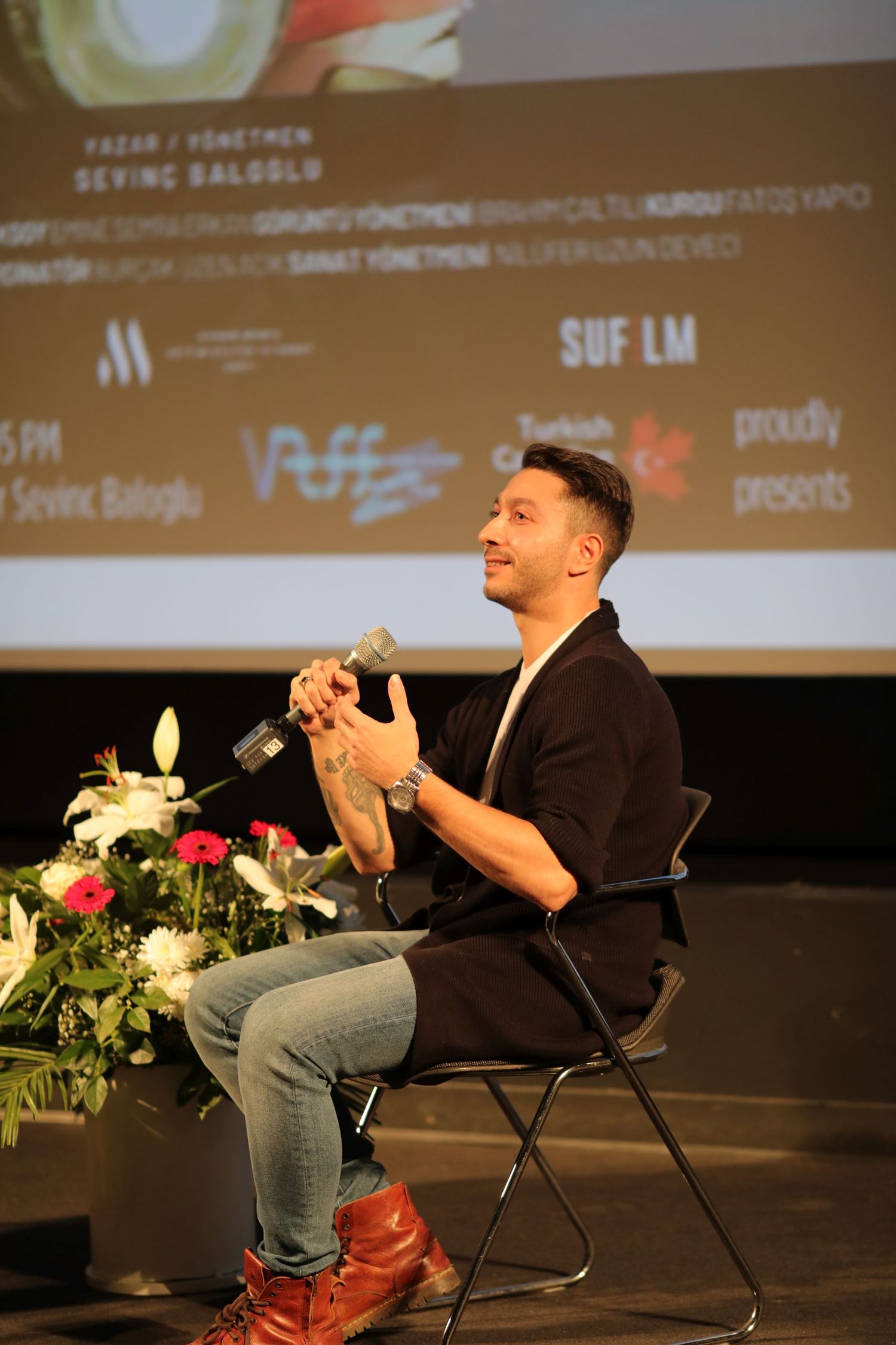 Speaker sits on stage holding a microphone during a presentation with projector screen and flowers in background.