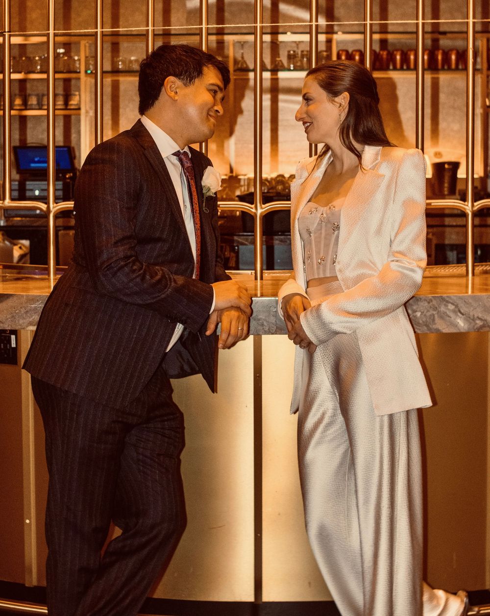 Elegant wedding couple sharing a laugh at a golden bar, bride in white suit, groom in pinstripe.