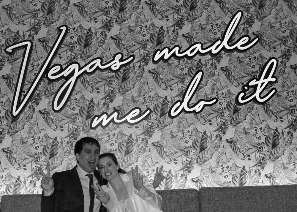 Black and white photo of newlyweds under Vegas Made Me Do It neon sign at wedding reception.
