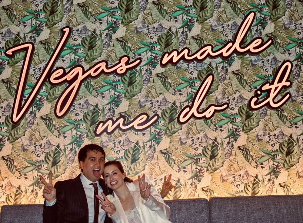 Cheerful couple celebrating under Vegas Made Me Do It neon sign with vibrant tropical wallpaper.