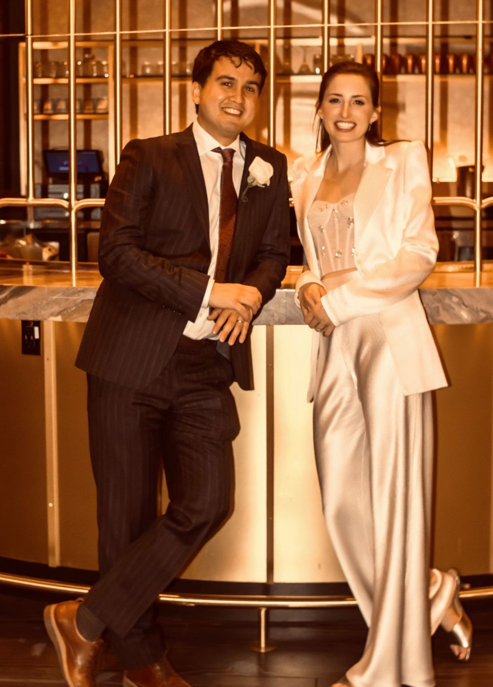 Smiling newlyweds posing at golden bar, groom in dark pinstripe suit, bride in white blazer set.