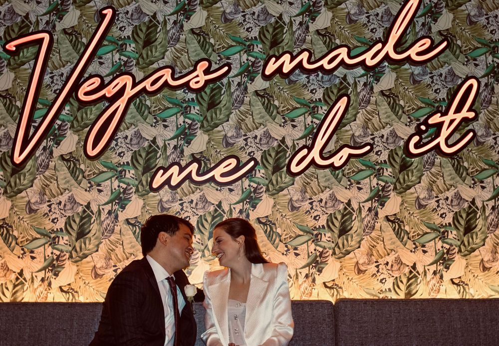 Newlyweds smiling under colorful Vegas Made Me Do It neon sign with floral wallpaper backdrop.