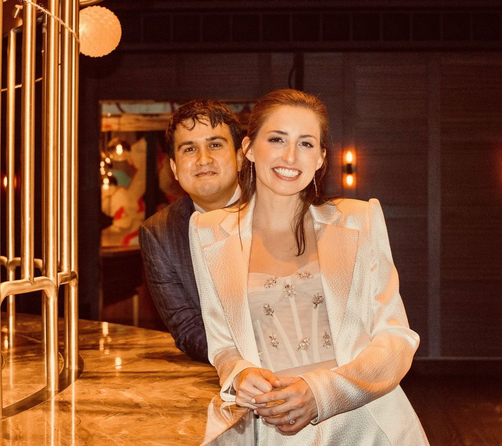 Smiling bride in white blazer poses with groom in blue suit at elegant venue.