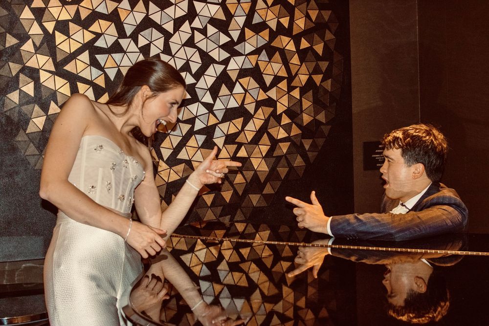 Groom playfully points at laughing bride in front of golden geometric wall decor.