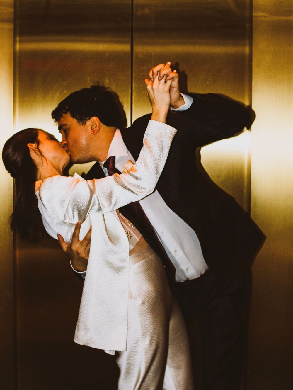 Newlyweds share passionate kiss while holding hands in golden elevator.