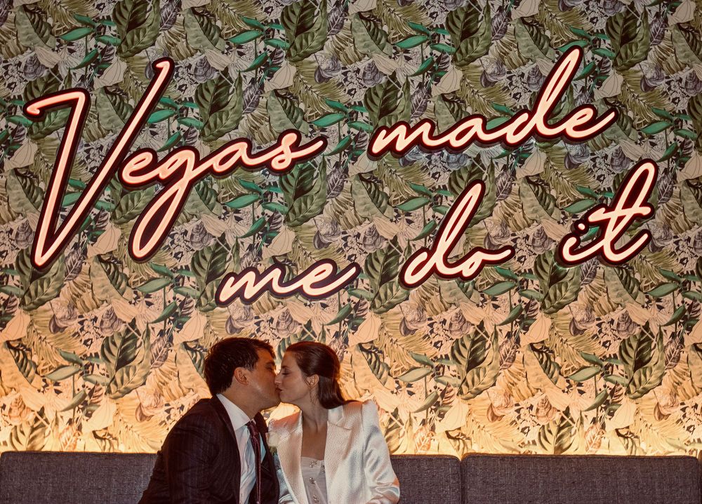 Newlyweds kissing under a glowing Vegas Made Me Do It neon sign with floral wallpaper backdrop.