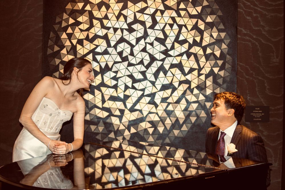 Bride in strapless gown and groom in dark suit laughing together beside a geometric mosaic wall.