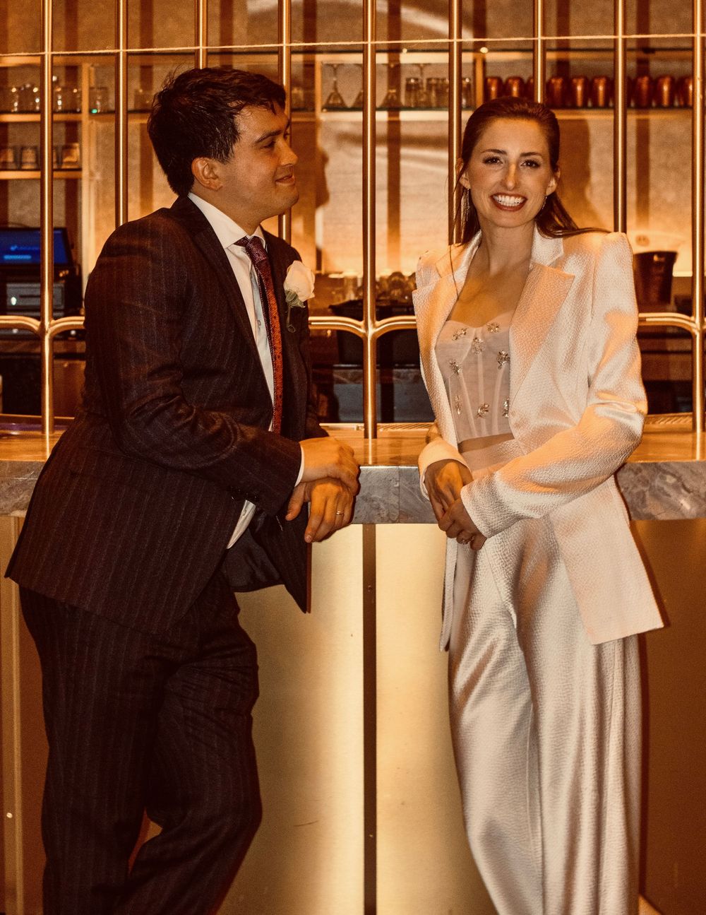 Smiling bride in white blazer and corset top posing with groom in pinstripe suit at a bar.