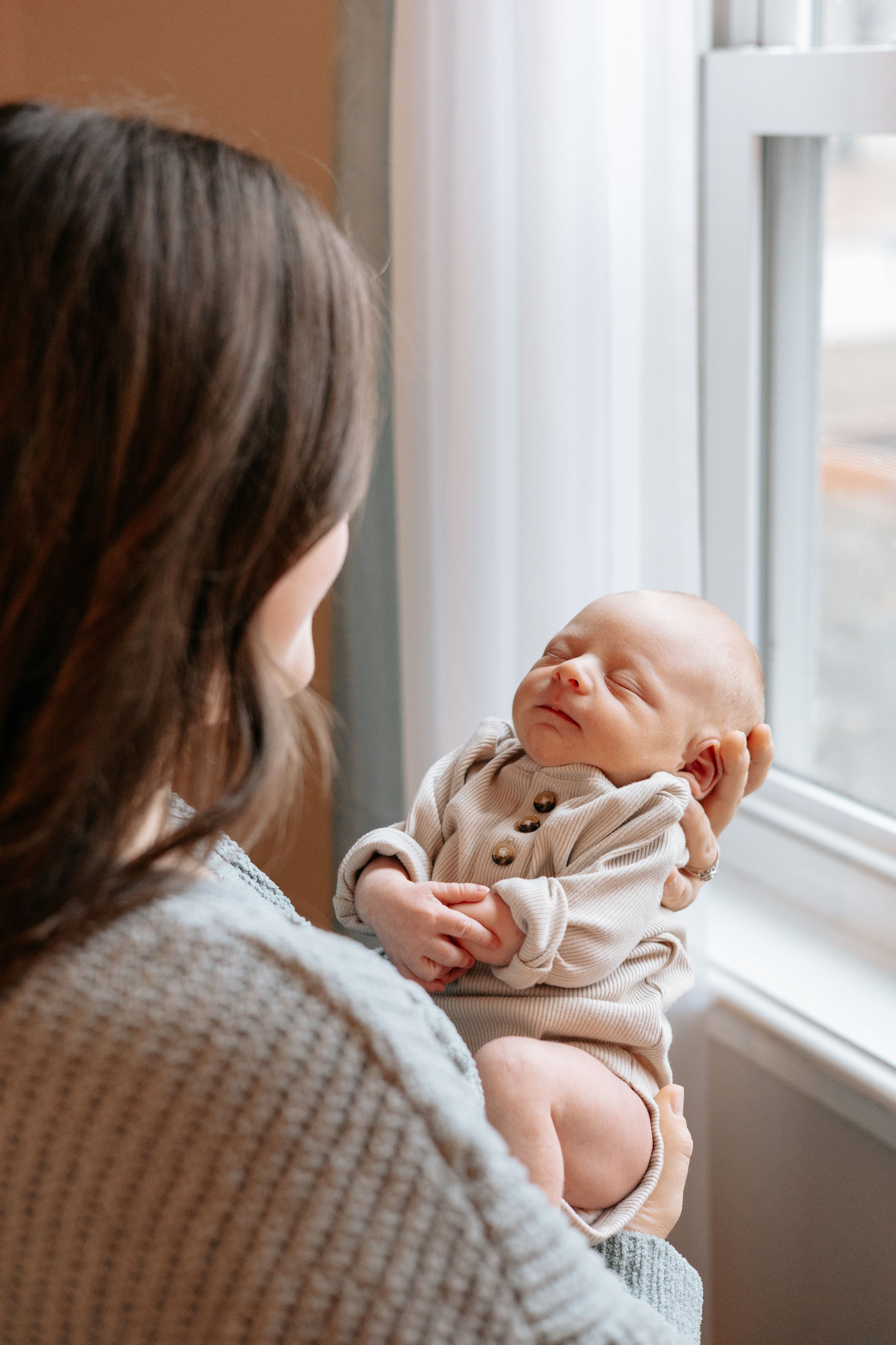 Tender moment by window as parent cradles newborn in soft natural light.
