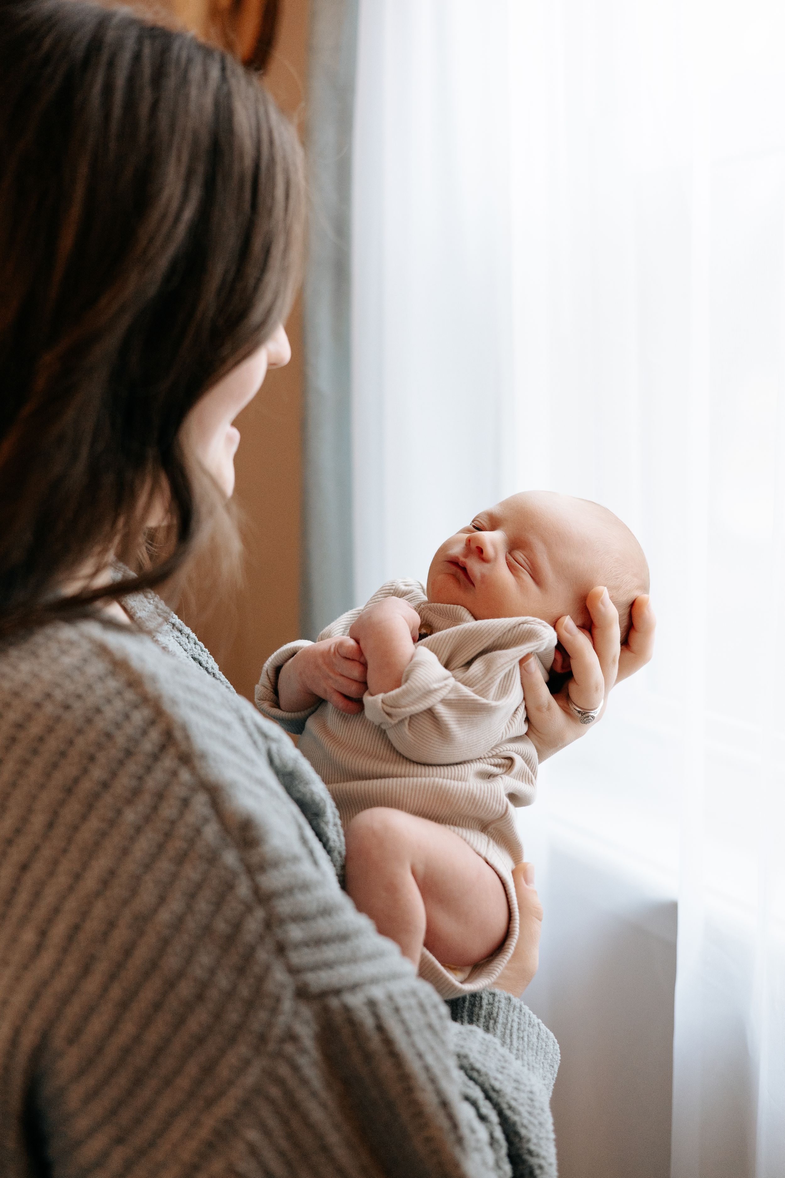 Tender moment between parent and newborn baby in soft natural window light wearing cozy knit sweater.