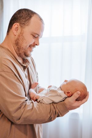 Parent in beige sweater tenderly holding and interacting with newborn baby near window.