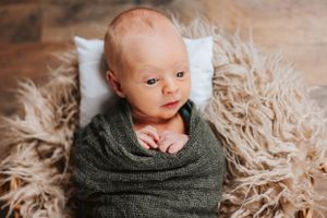 Infant wrapped in dark green swaddle resting on fluffy beige blanket.