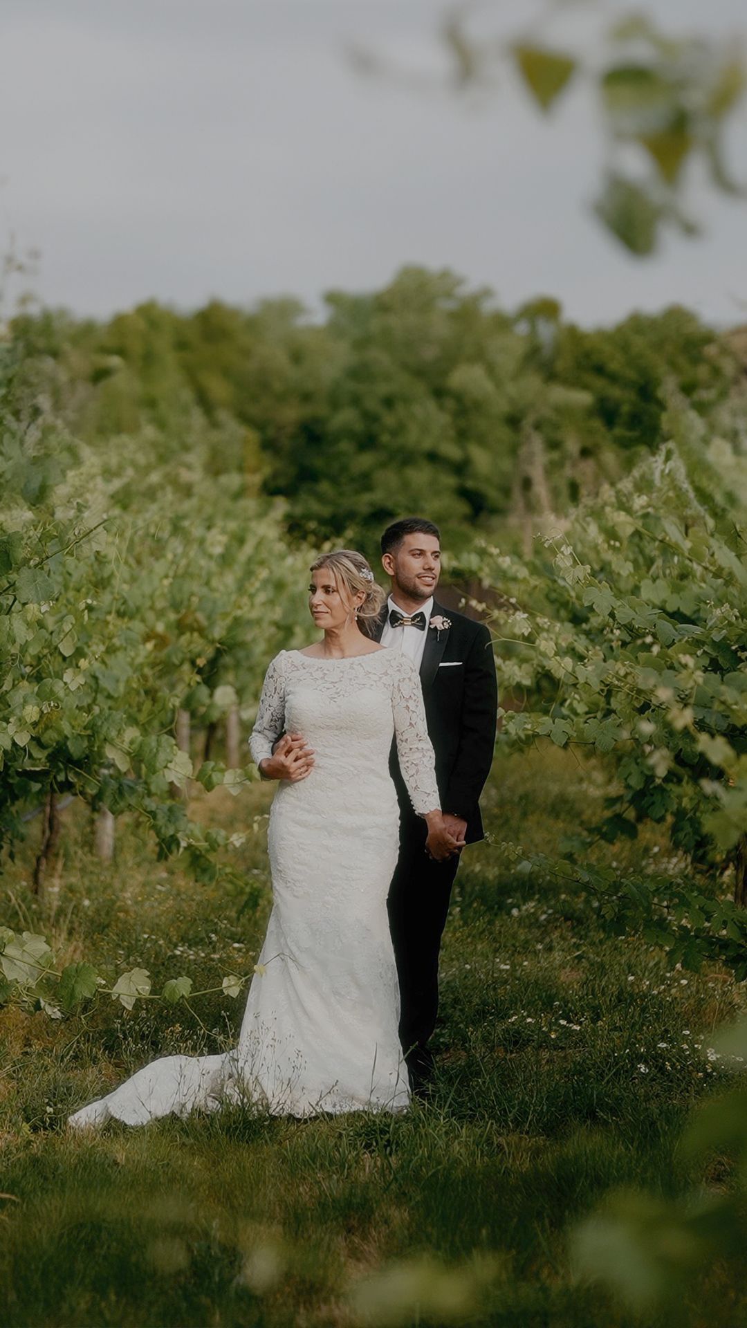 A bride in a lace gown stands with her groom in a vineyard during a romantic outdoor wedding photoshoot.