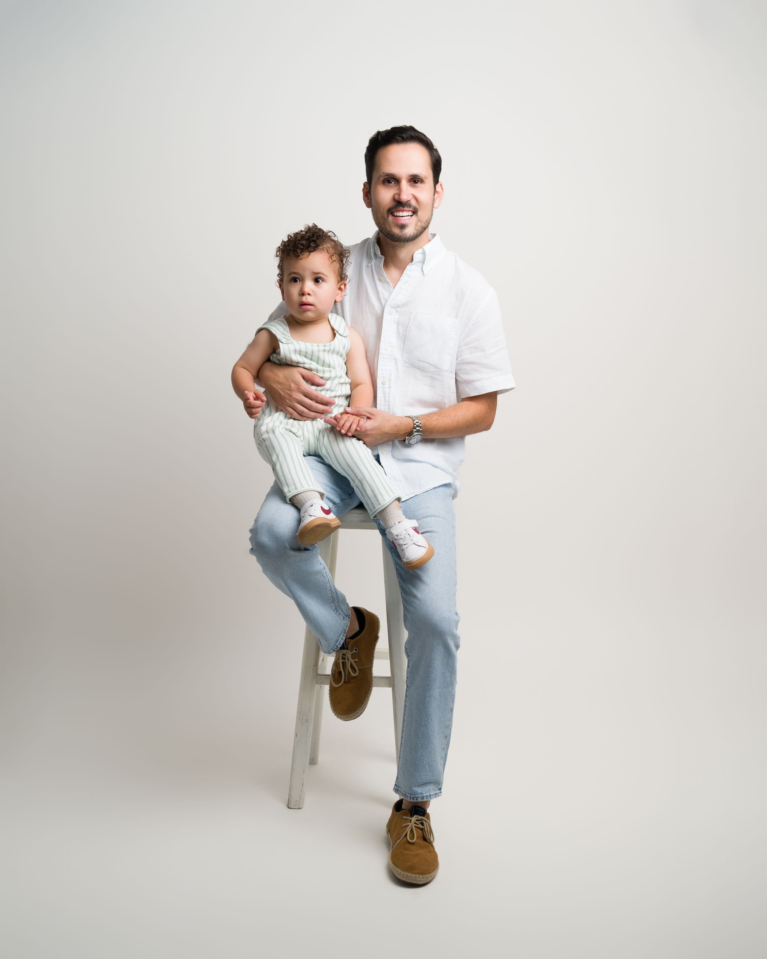 Smiling father in white shirt holds young toddler while sitting on stool in studio.