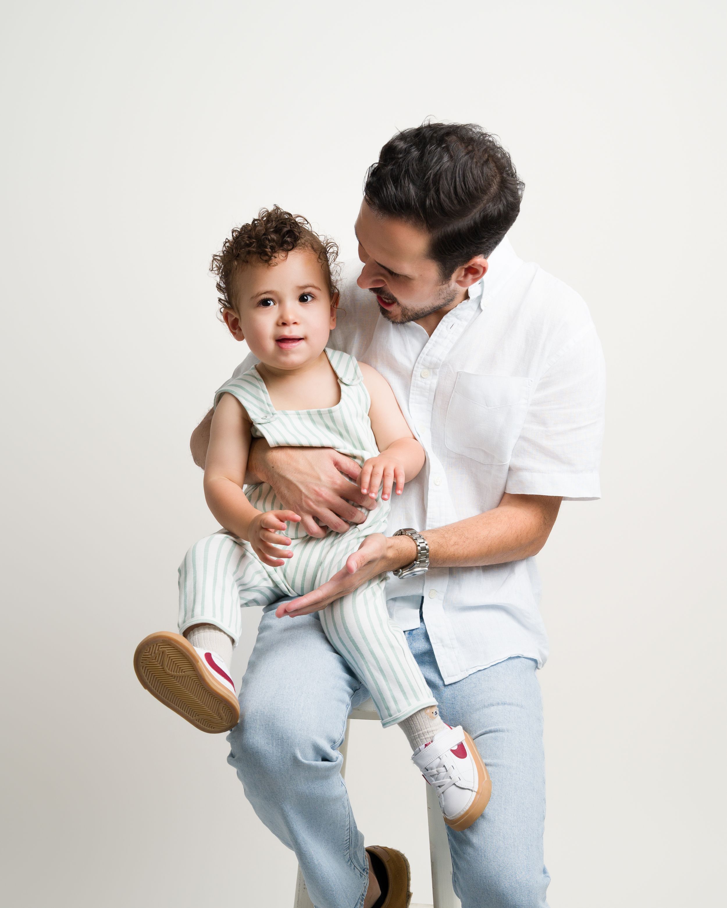Father holding toddler boy on white stool in studio portrait.
