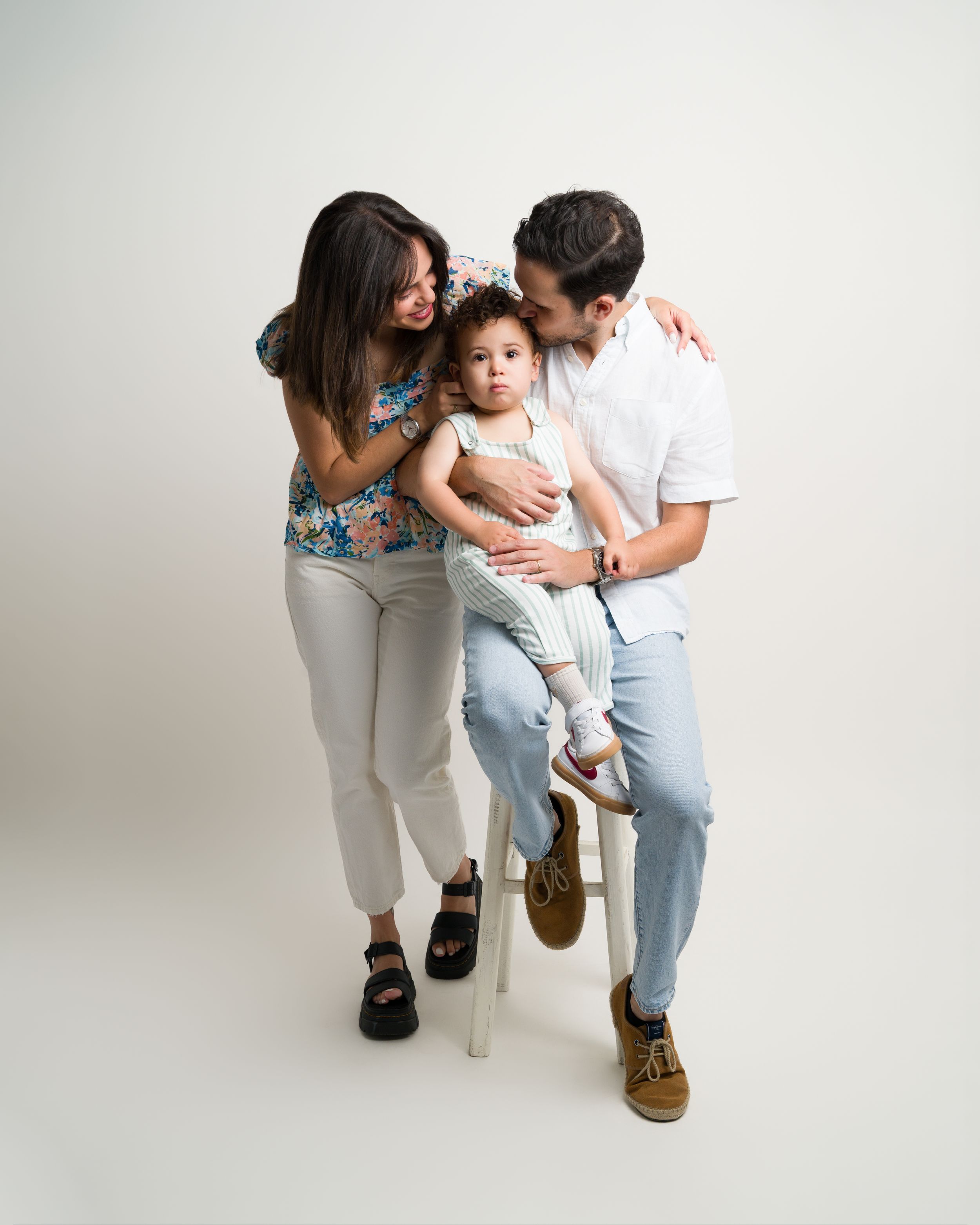 Family portrait of parents kissing toddler boy on white stool.