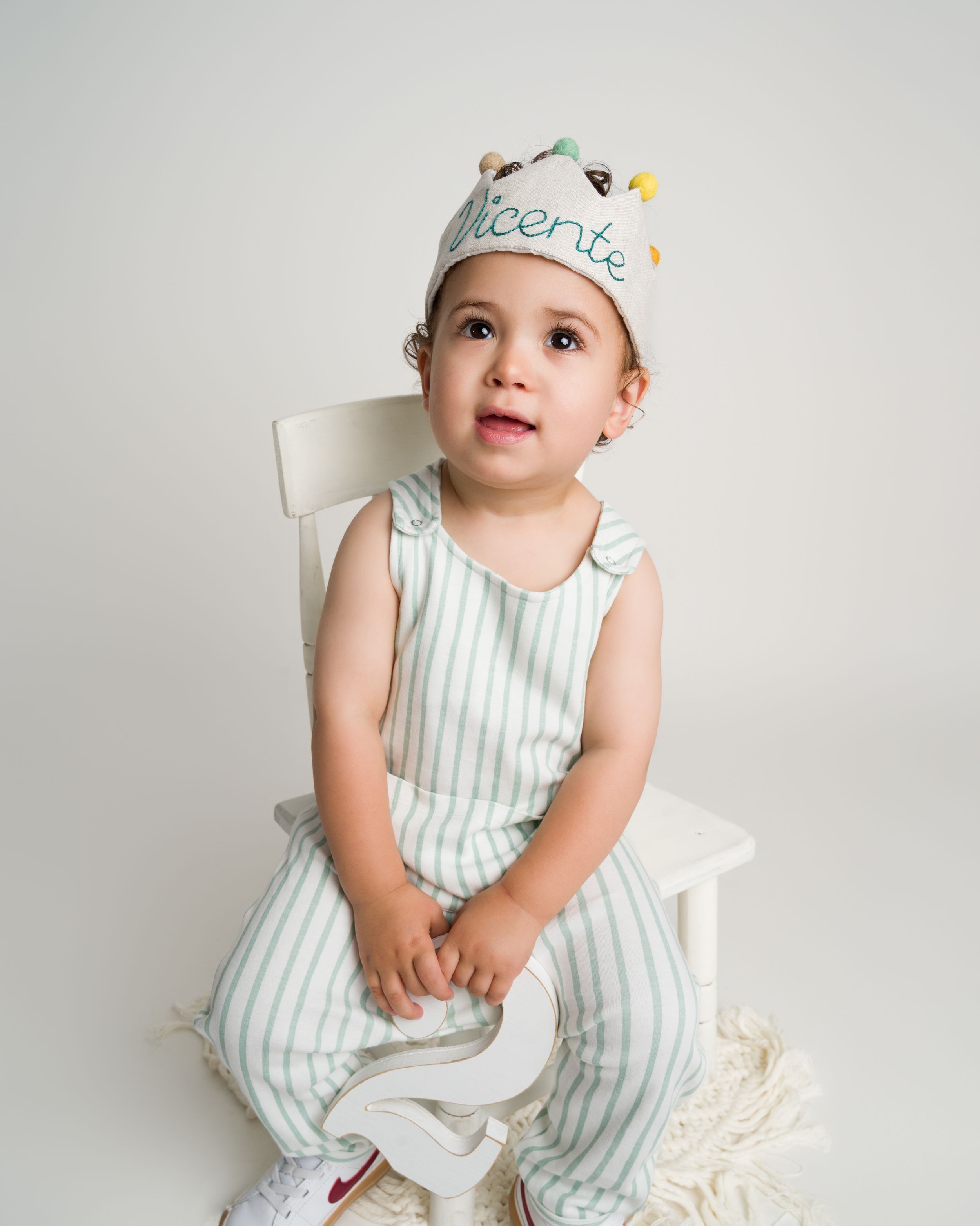 Toddler boy Vicente in birthday crown sitting on white chair.