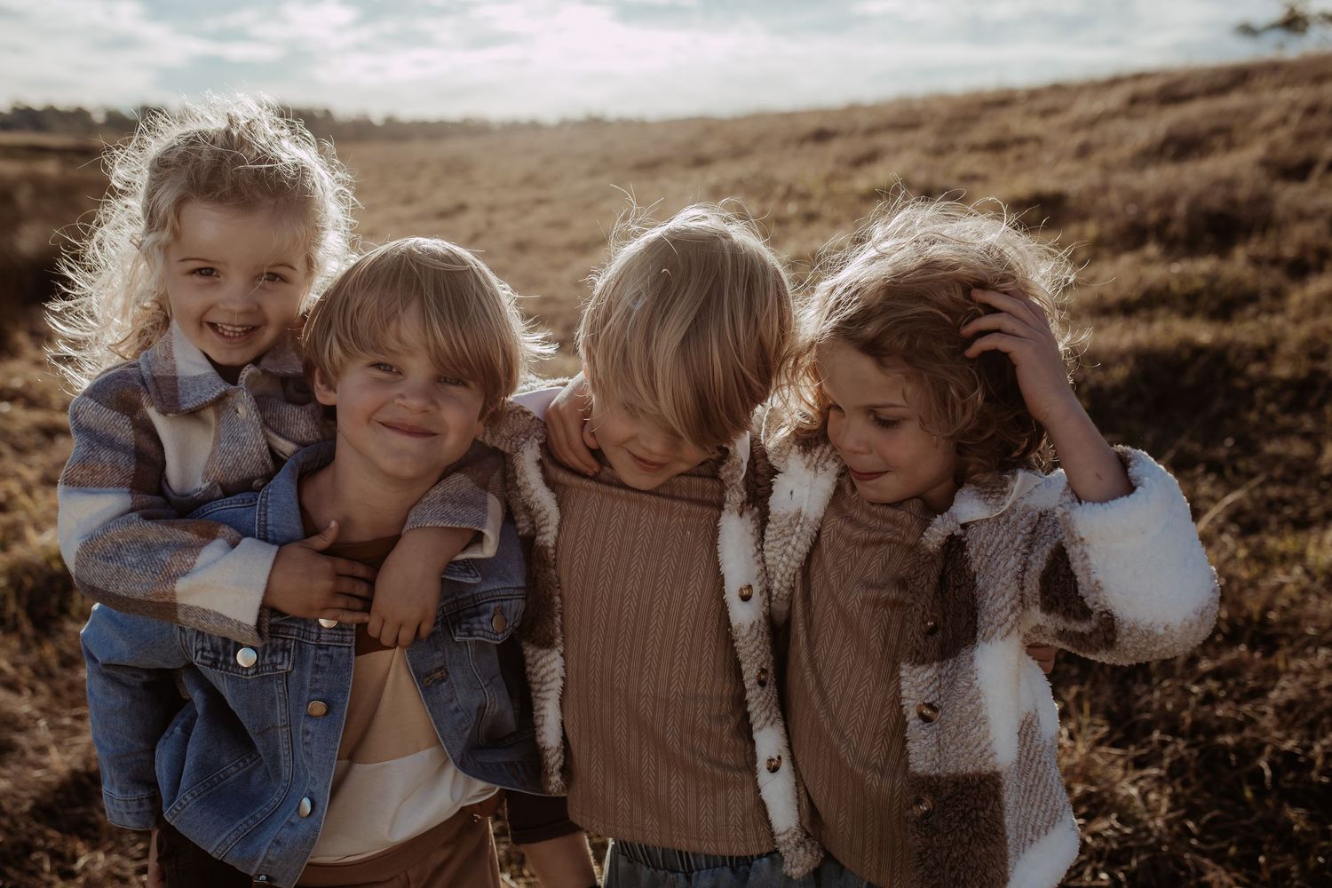 Four playful children laughing and huddling together in a sunny field during golden hour.