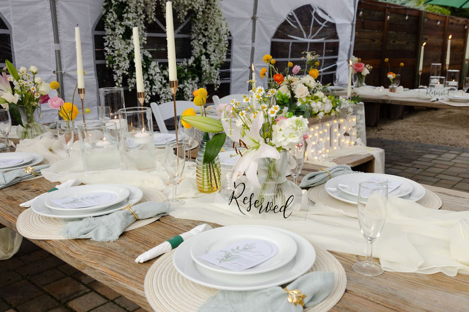 Elegant outdoor wedding reception tables set with white linens, floral centerpieces and rustic wooden chairs.