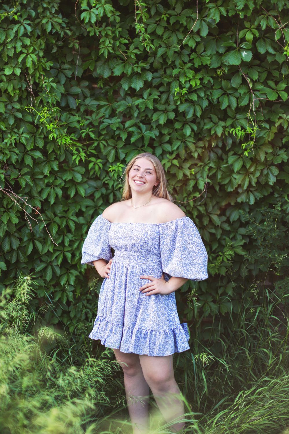 High school senior wearing a blue and white floral peplum top poses in front of lush green foliage.