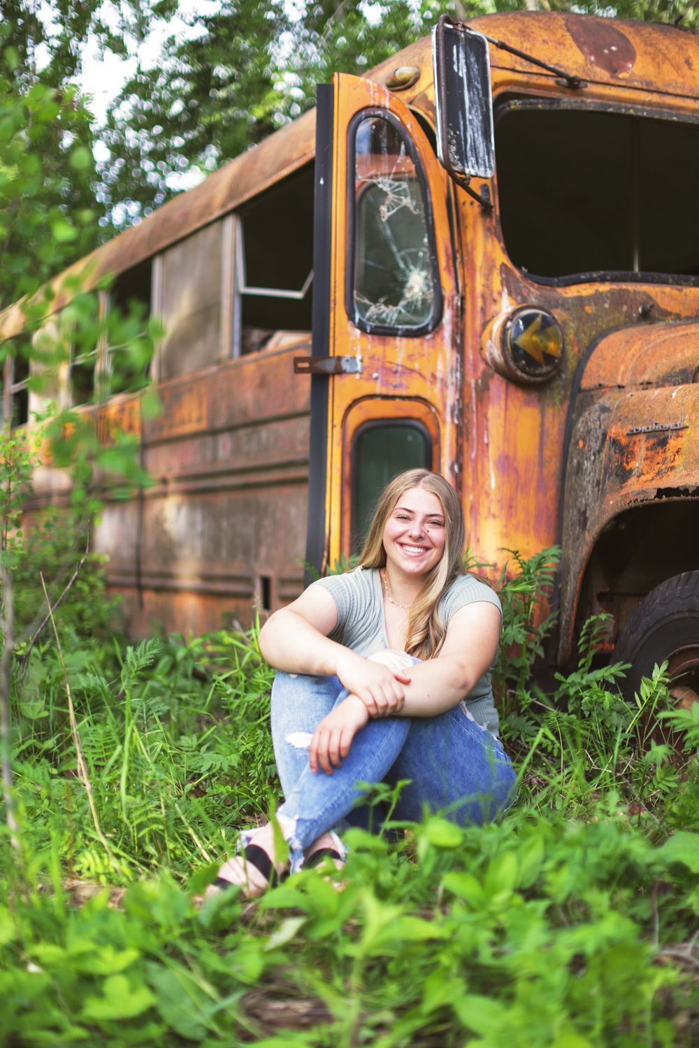 A rustic Senior photo posed beside an old abandoned orange school bus in overgrown grass.