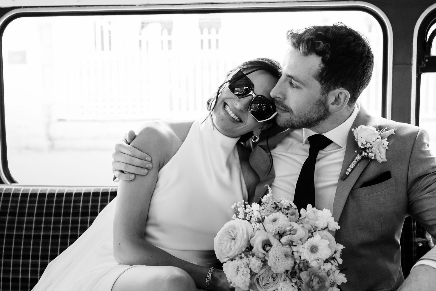 A newlywed couple shares a romantic kiss while sitting in a vintage van with white floral bouquet decorations.