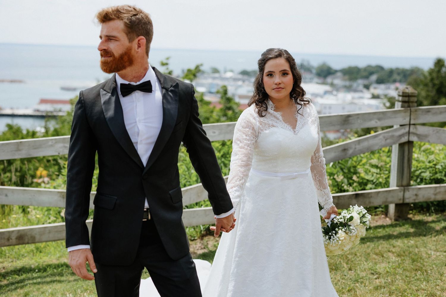 A couple in formal wedding attire walk together and hold hands with the view of Mackinac Island below
