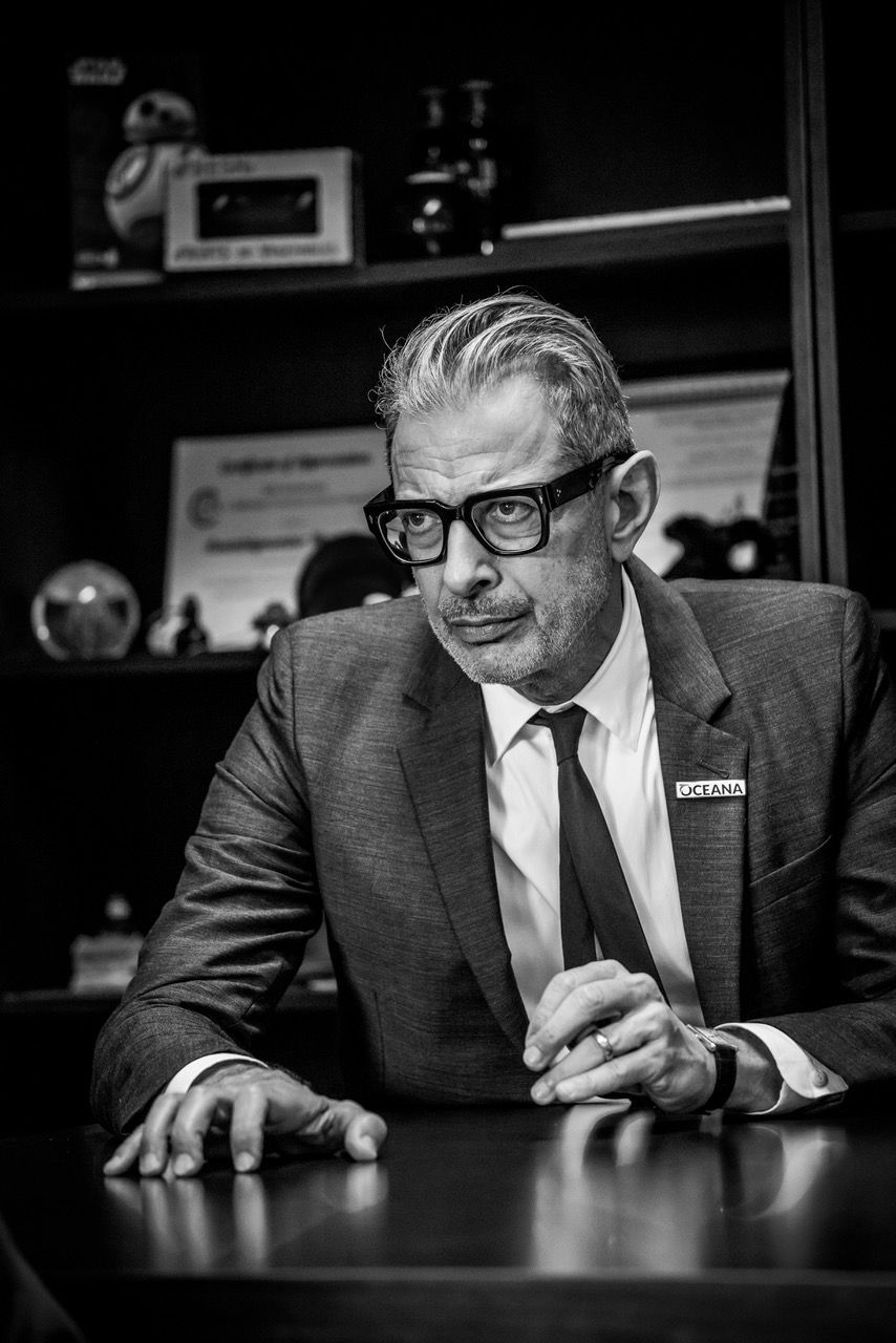 Black and white portrait of a man in suit and glasses sitting at a desk with shelves in background.