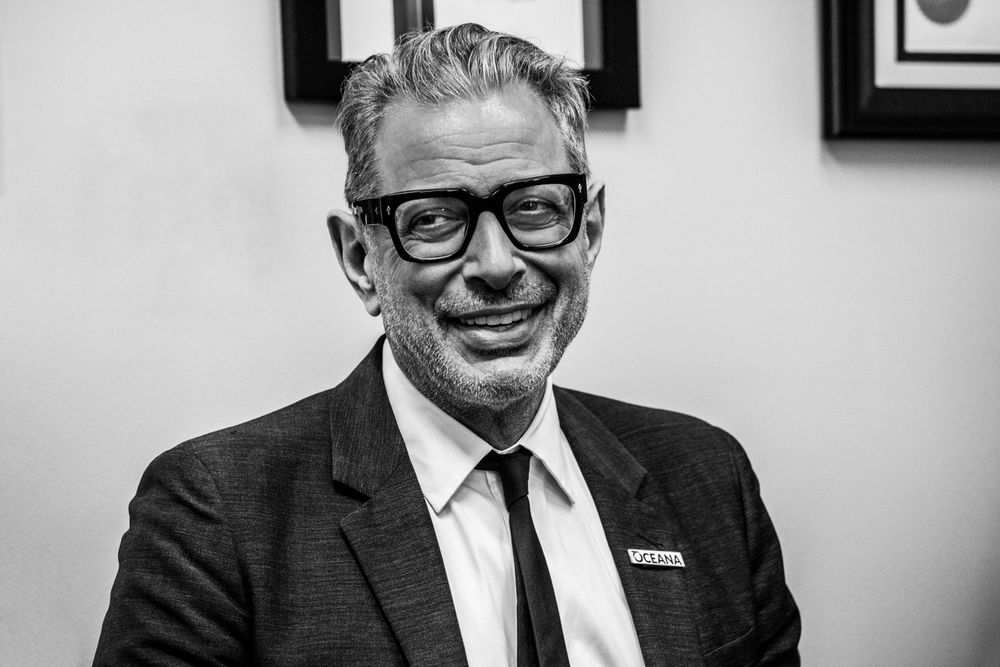 Black and white portrait of a smiling older man wearing thick-rimmed glasses and a suit jacket.
