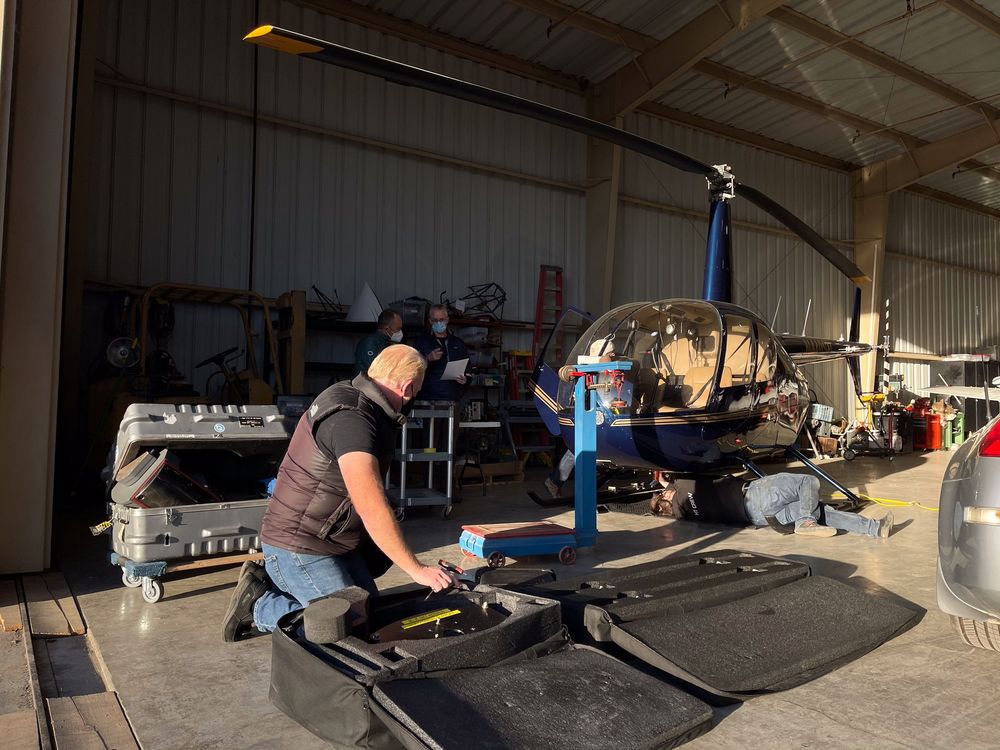 Mechanic performing maintenance on a helicopter inside an aviation hangar with tools and equipment nearby.