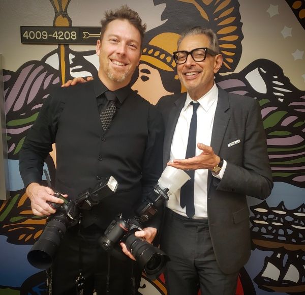 Two smiling men pose together, one holding professional cameras, in front of a colorful mural backdrop.