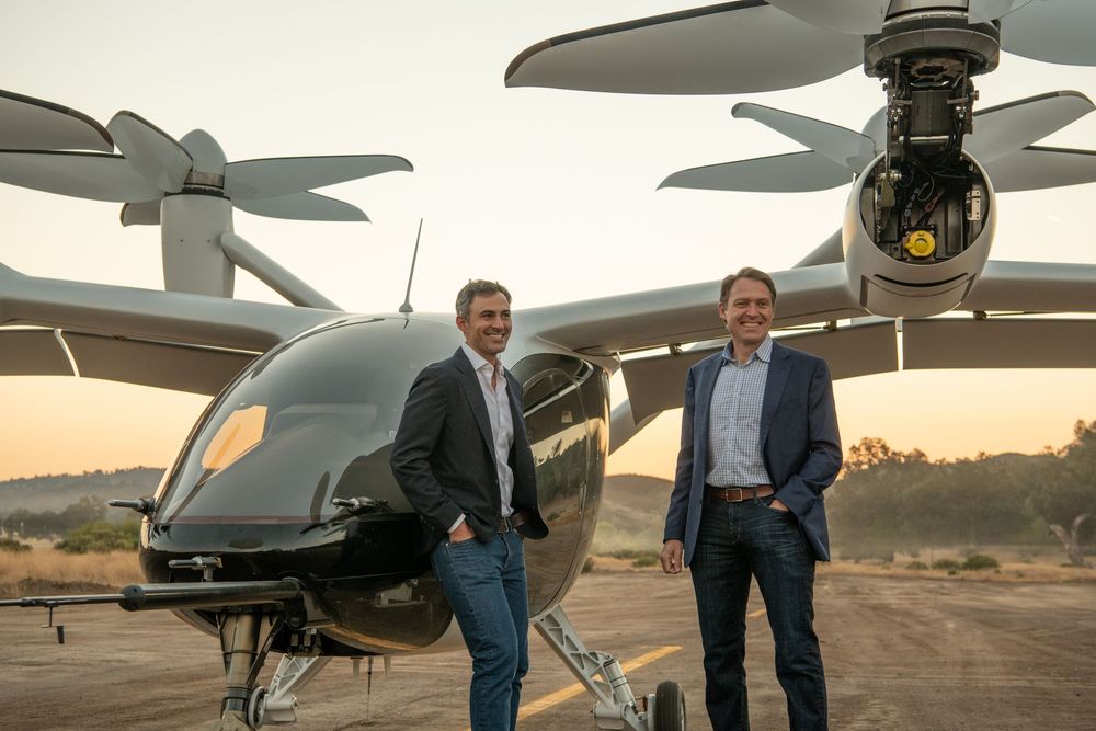 Two men smile standing in front of a modern electric aircraft on a runway at sunset.