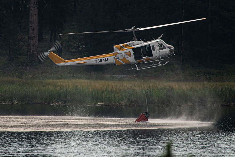 A yellow and white helicopter with registration N354M skims over a lake, filling a water bucket for firefighting operations.