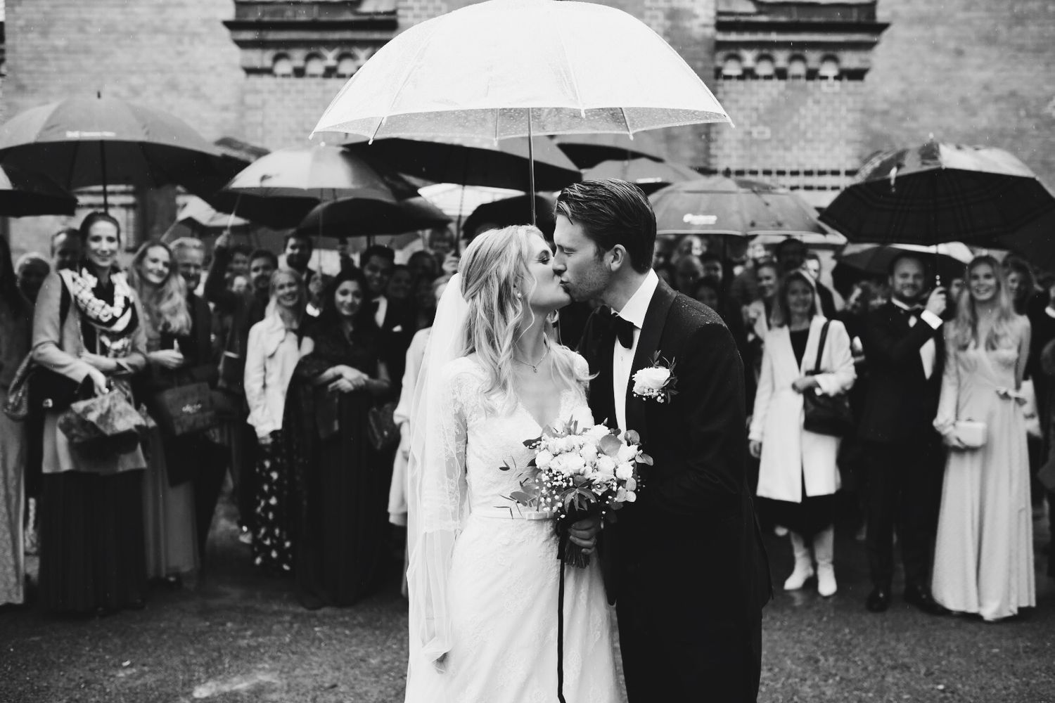 A rainy wedding scene with guests holding black umbrellas around the couple in black and white.