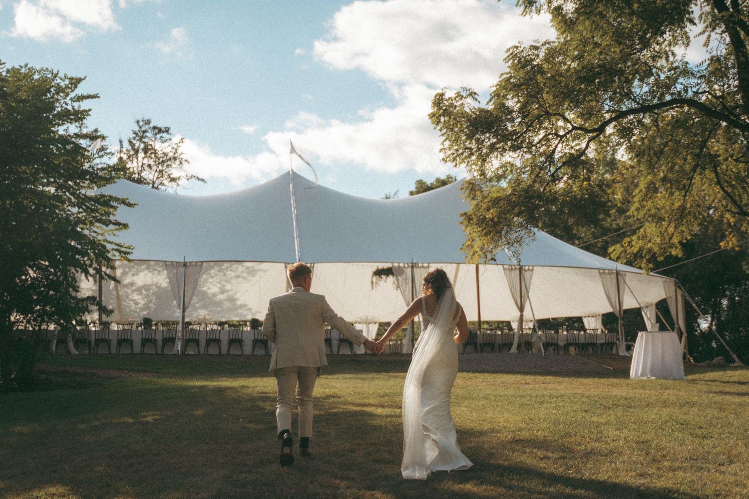 Couple in wedding attire strolling across lush grass toward an elegant white sailcloth reception tent.