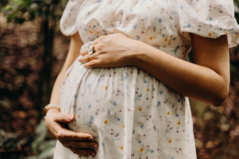 A person in a white floral dress holds their pregnant belly while standing in a natural outdoor setting.