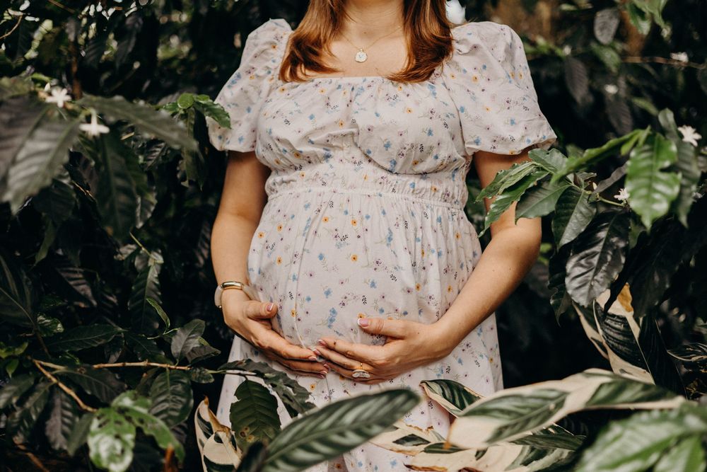 A pregnant woman in a floral dress poses in a garden setting with lush green foliage surrounding her.