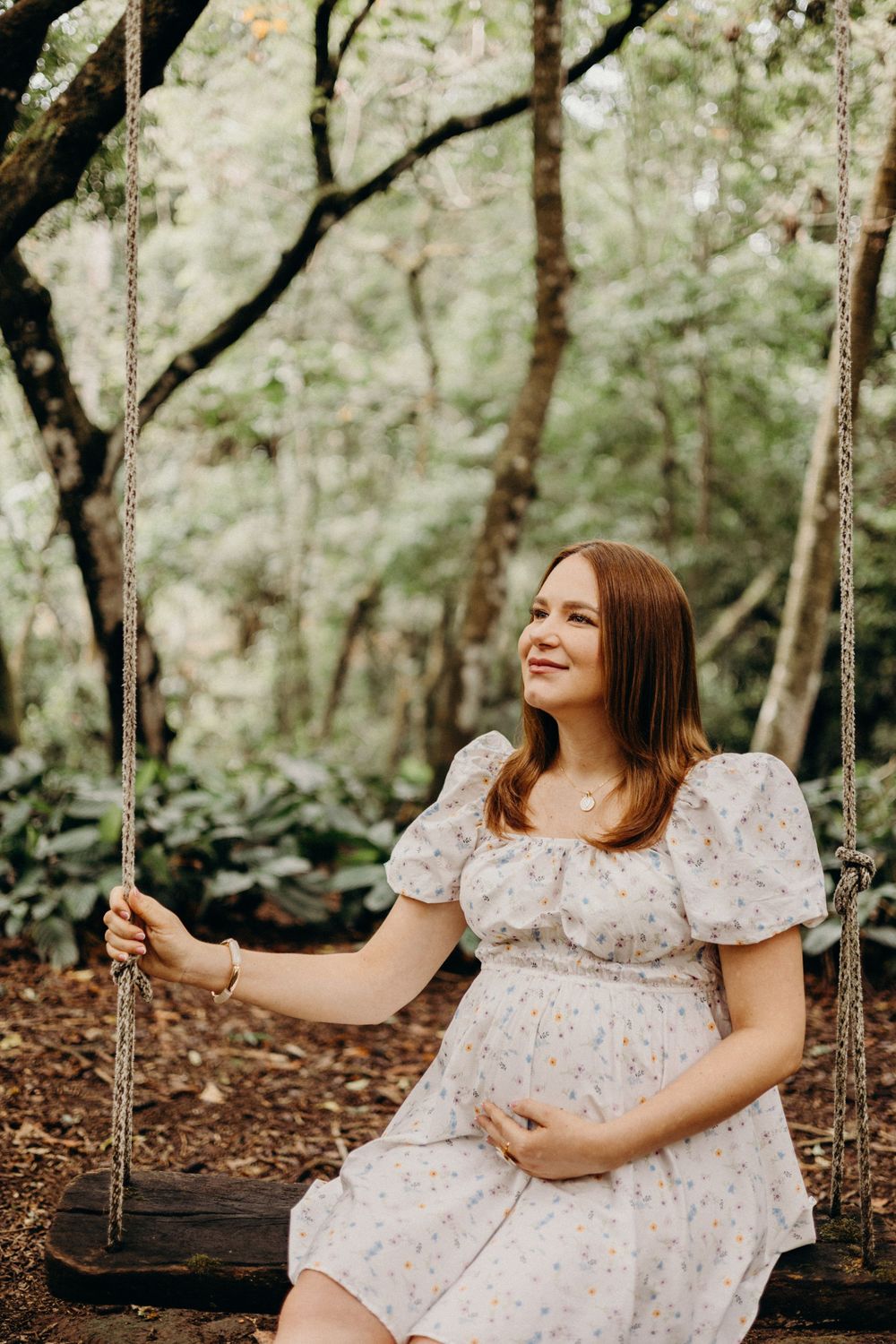 Woman in floral dress sitting on tree swing in enchanted forest setting during outdoor photoshoot.