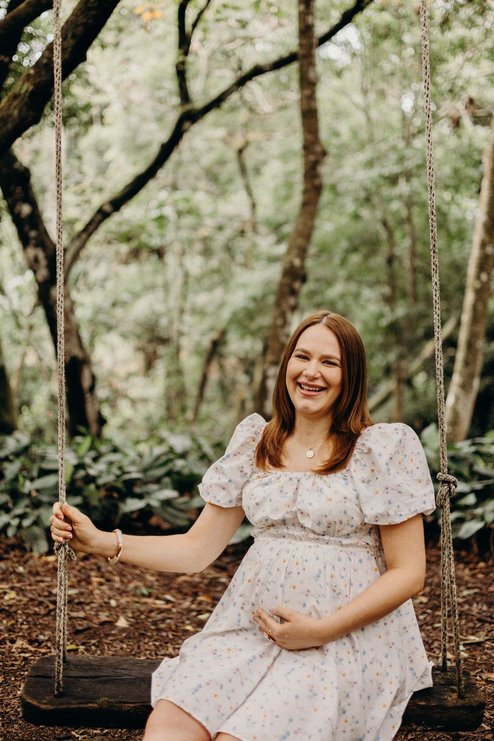 A floral dress-clad figure sits on a wooden swing in a peaceful wooded setting.