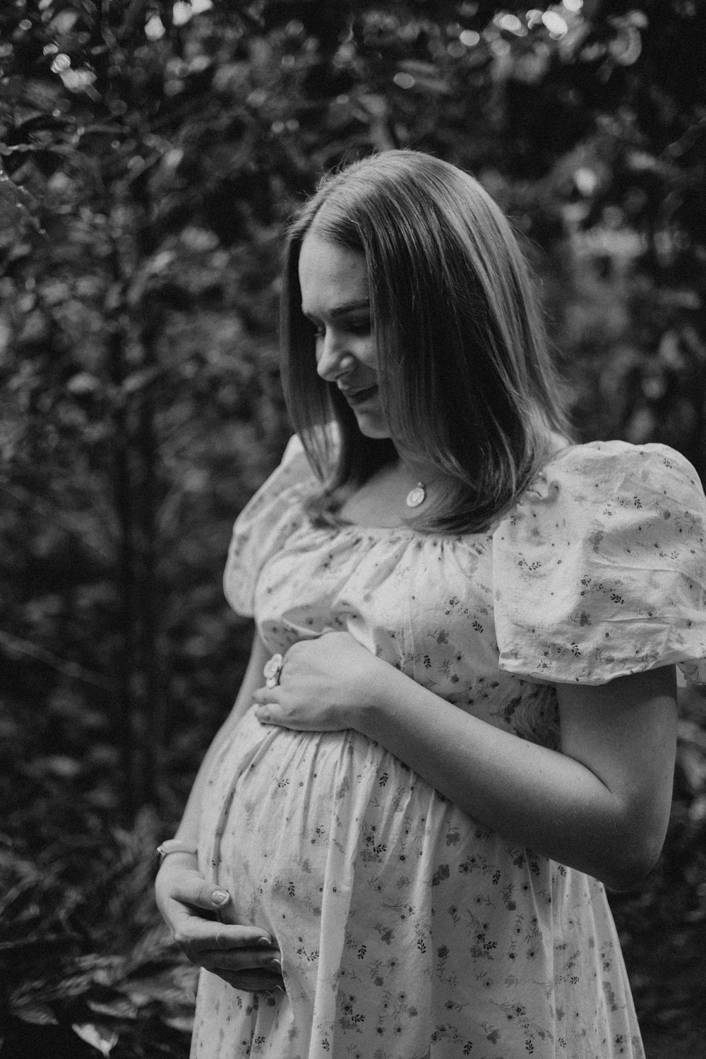 Black and white portrait of an expectant mother in a floral dress tenderly cradling her baby bump in a garden setting.